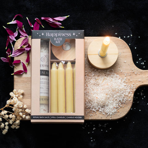 Lifestyle image of happiness bath kit, gift box contains candles, candle holder and bath salts, presented in a beautiful gift box. The image includes a display board and flower petals.