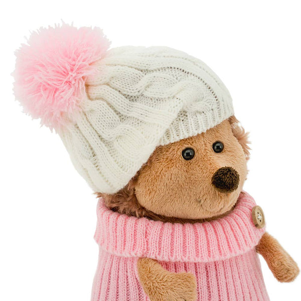 Side facing studio shot of a plush hedgehog soft toy, dressed in a pink jumper and a white wolley hat with a pink pom pom.